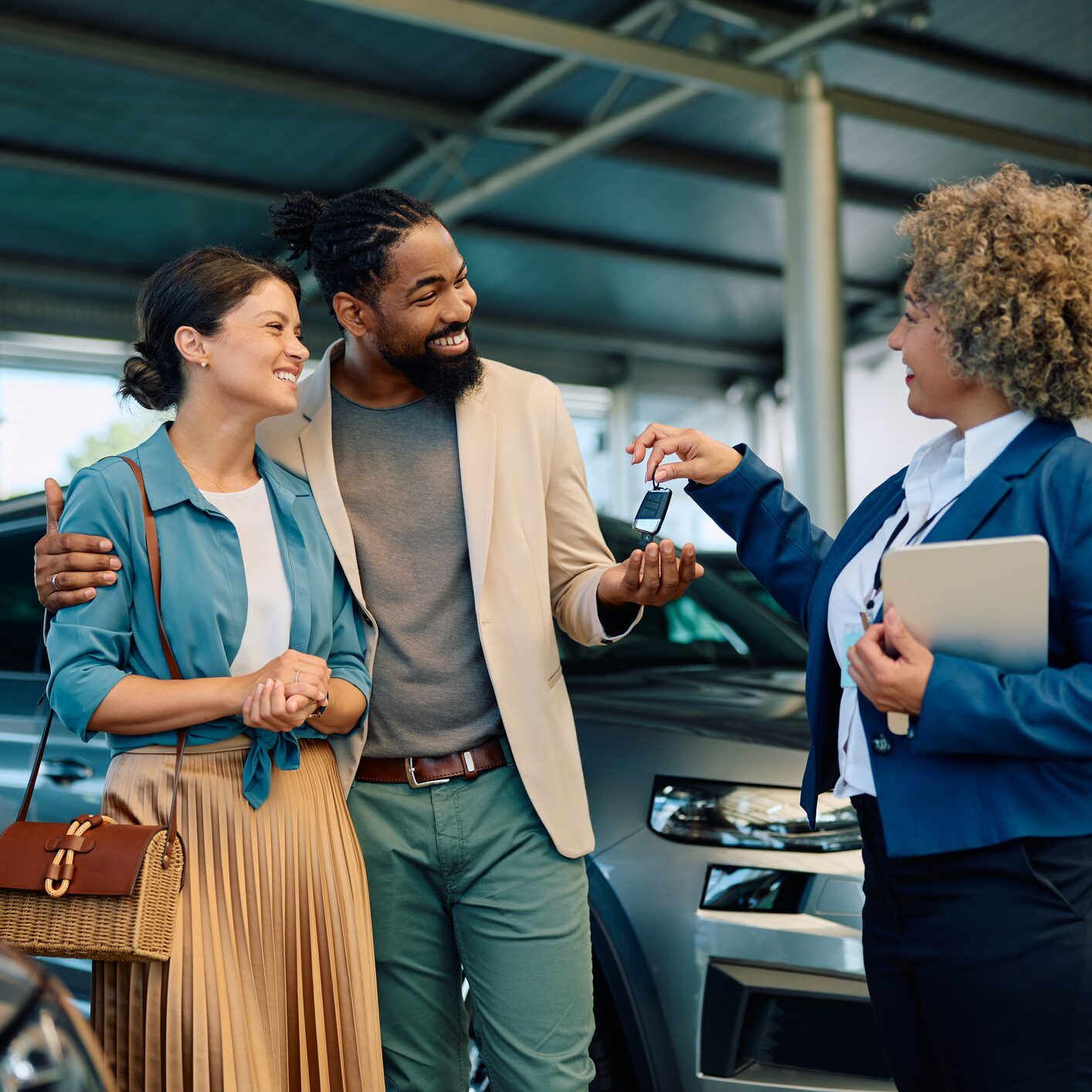 Happy multiracial couple receiving keys of their new car from saleswoman in showroom.